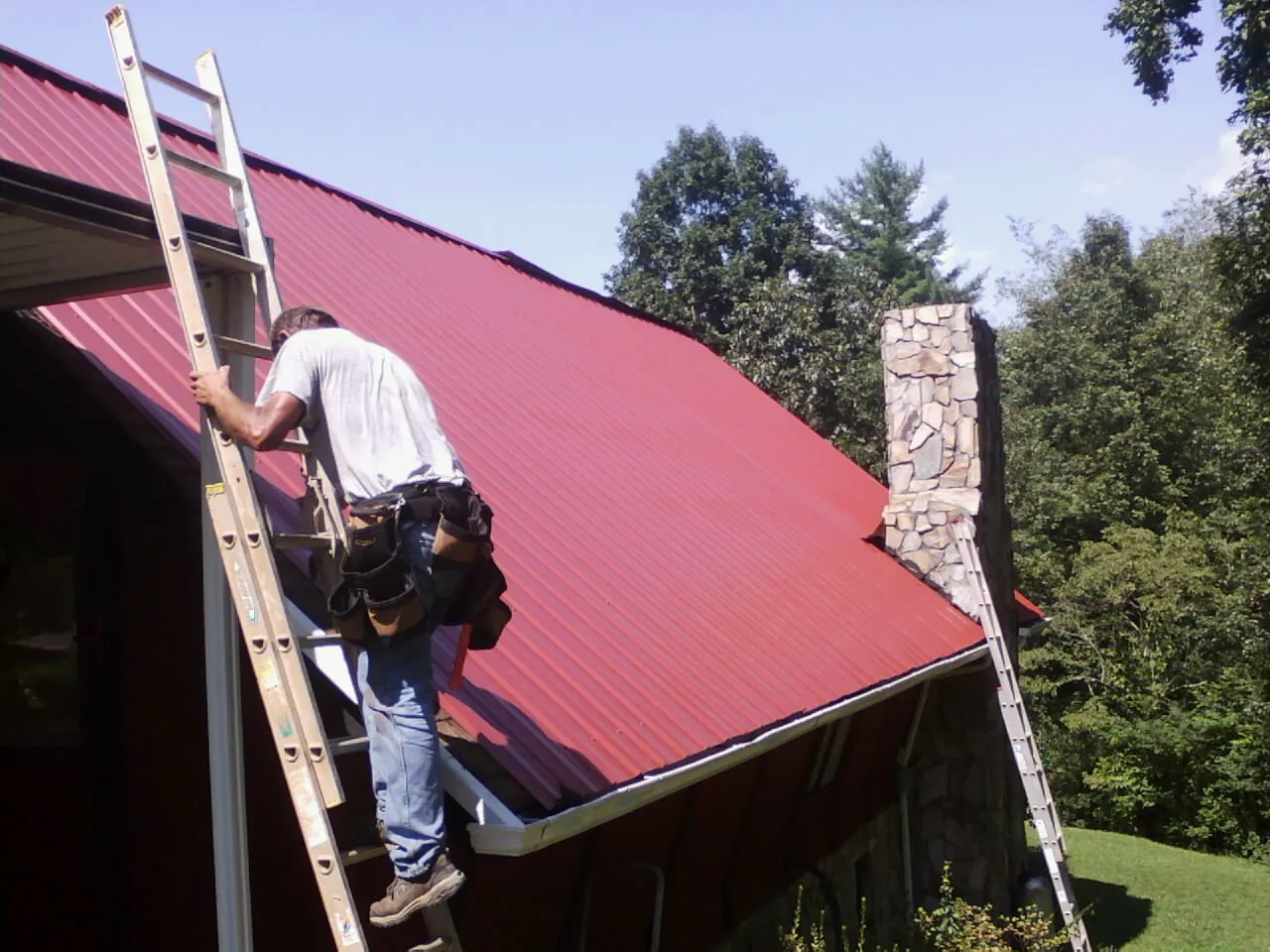 Roofer on a ladder beside a red metal roof and stone chimney — Soffit & Fascia Repair by Roofing Brothers in Cleburne